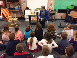 Ellen Rumsey with a group of children reading the story book. There are about 15 children sitting crossed legged listening quietly to Ellen.