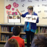 Image of Ellen Rumsey reading to a group of young children. She is wearing a Blue Sweater and Black Pants. She is pointing to a storyboard.