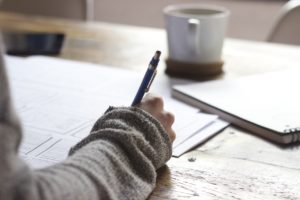 A person writing in a diary at a desk with coffee in the background. The person's hand is writing with a pen in their right hand. 